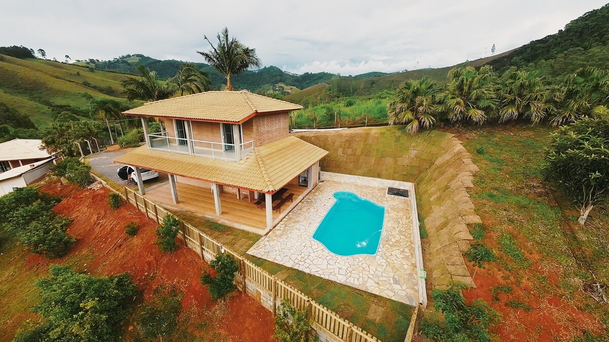 An aerial view reveals a two-story house surrounded by lush greenery. A private pool with clear blue water is situated beside a spacious wooden deck, enhancing the outdoor appeal. The property is fenced, providing privacy and a serene atmosphere, ideal for family relaxation.