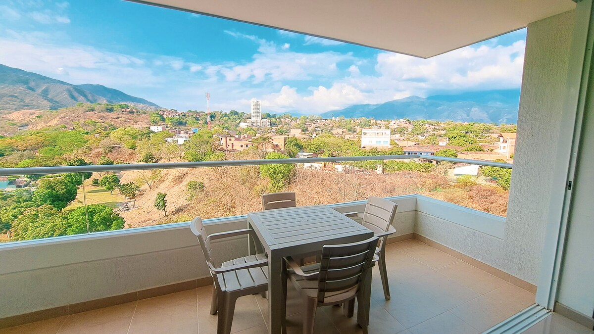 A spacious balcony features a dining table surrounded by several chairs, offering expansive views of the surrounding landscape and mountains. The open area allows sunlight to fill the space, providing a serene setting for outdoor relaxation.