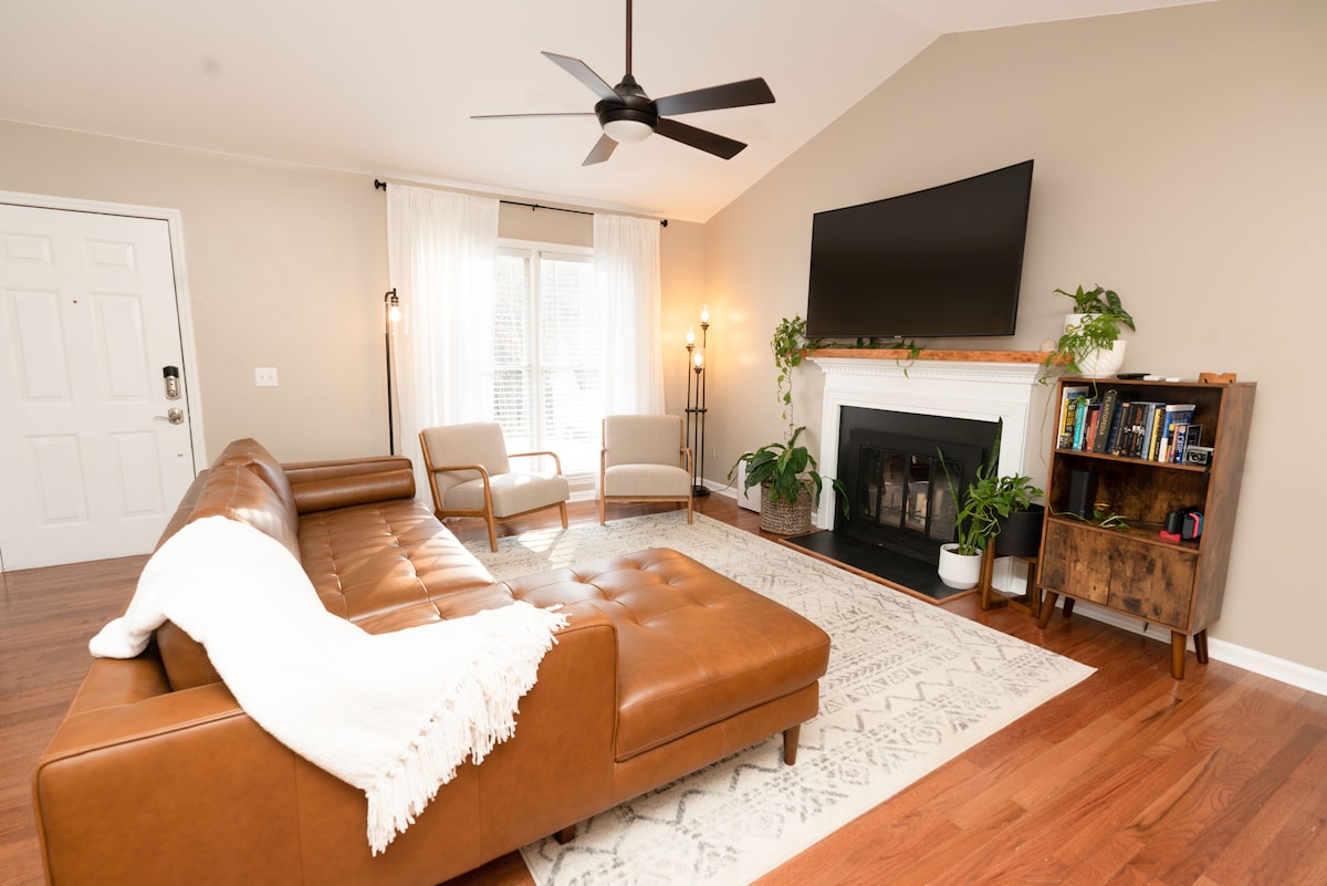 A spacious living area features a leather sectional sofa accompanied by two armchairs. A flat-screen television is mounted above a modern fireplace, with decorative plants on either side. A bookcase holds a variety of books, and light flows through the large window covered with sheer curtains.