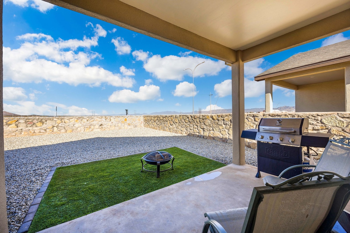 A covered patio area features a grill and seating arrangement with views of a spacious gravel outdoor space. Grass patches are visible along with a stone wall, providing a defined boundary for the area. The sky is bright with scattered clouds.