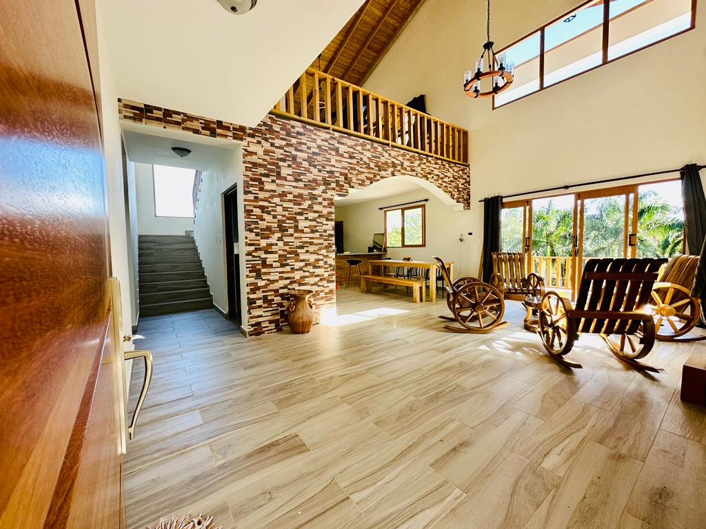 An expansive living area features a high ceiling with wooden beams and large windows that allow natural light to fill the space. Stone accents on the walls complement two wooden rocking chairs positioned to invite relaxation, while a dining table is visible in the background.