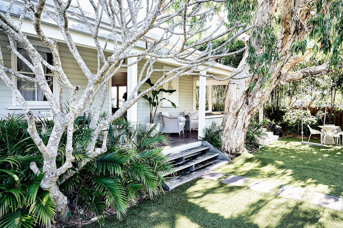 An outdoor view of the cottage reveals a wide porch with comfortable seating, framed by lush greenery and trees. The well-maintained yard features manicured grass and a stone pathway leading toward the entrance, creating a welcoming ambiance.