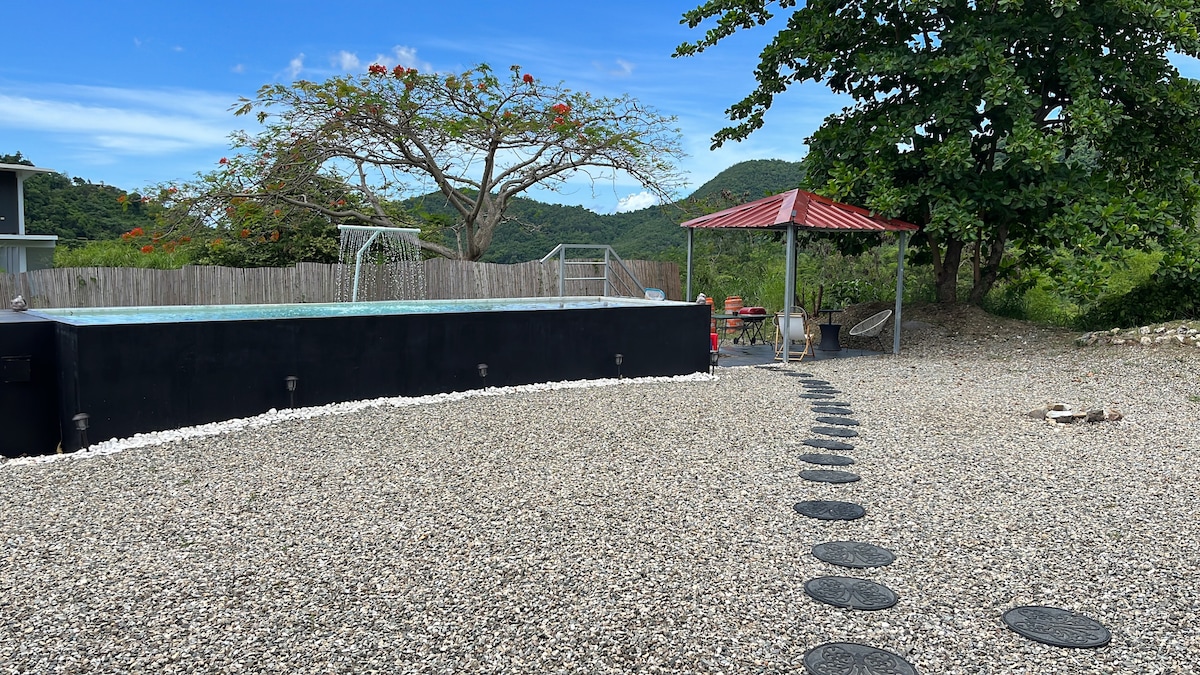 A private pool is set amidst a tranquil outdoor space, surrounded by a gravel pathway. Lounge chairs and a gazebo provide shaded seating, with lush green mountains visible in the background. A tree stands nearby, enhancing the natural ambiance.