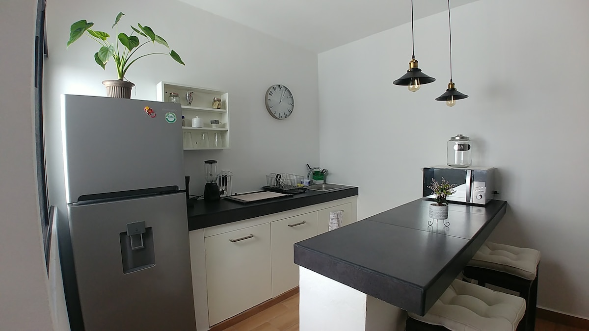 A modern kitchen is visible, featuring a sleek countertop and ample cabinetry. A refrigerator stands against one wall, complemented by a clock and coffee maker on the countertop. Three stools are arranged at a breakfast bar, along with a plant adding a touch of greenery.