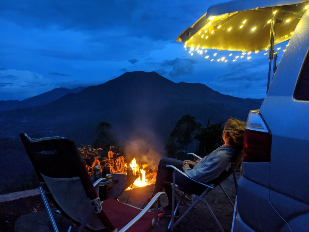A serene evening setting features a campervan parked next to a small campfire. Fairy lights adorn the canopy above, providing soft illumination. A figure is seen comfortably seated in a camping chair, with a backdrop of distant mountains under a twilight sky.