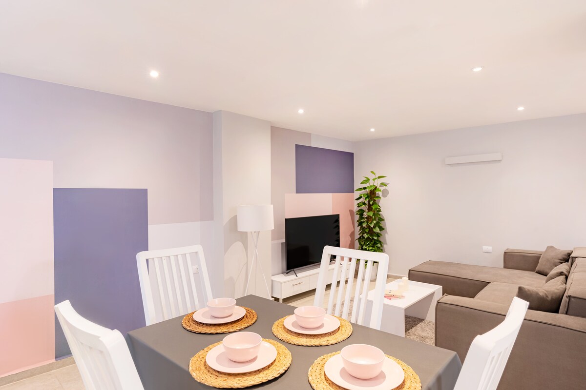 A dining area is seen with a table set for four, featuring pale pink dishes on woven placemats. Light grey walls with abstract pastel shapes provide a modern backdrop. A cozy seating arrangement with a large sofa is visible to the right, along with a smart TV on a white console.