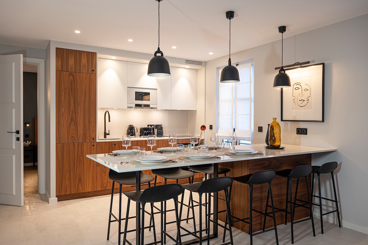 A modern kitchen features sleek cabinetry with wood accents, complemented by a spacious island. Four high-backed stools are arranged around the island. Pendant lights hang above, illuminating the countertop. Appliances, including a microwave and coffee maker, are integrated seamlessly into the design.