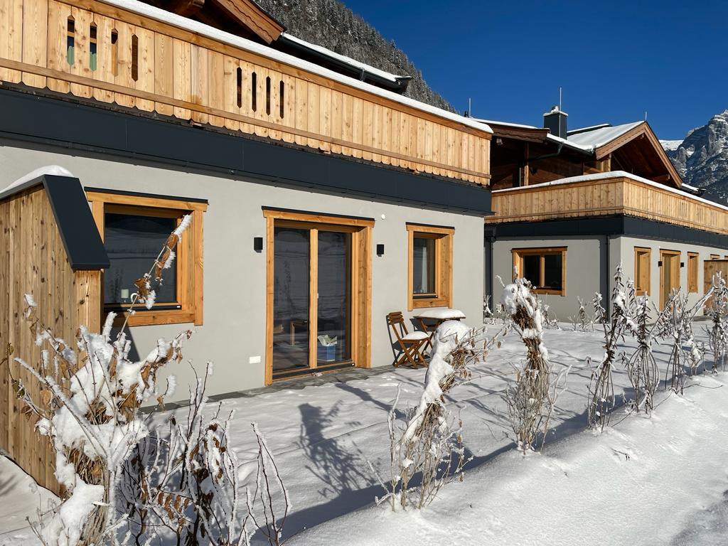 A modern building with wooden accents is framed by snow-covered terrain. Large windows are present, allowing natural light to enter. A small wooden table and chairs are placed outside, surrounded by white snow and foliage, with distant mountains visible under a clear blue sky.