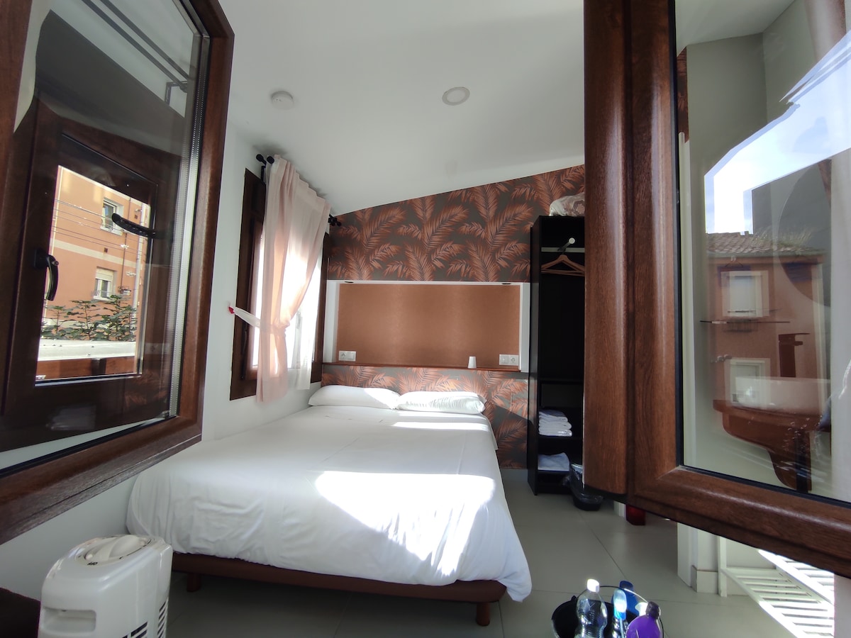 A well-lit individual room features a spacious bed dressed in white linens. A decorative wall with a floral pattern adds visual interest. A wardrobe stands nearby, while large windows allow natural light to fill the space, offering views of the outside.
