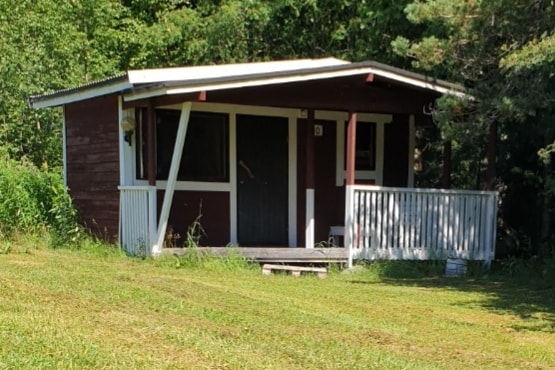 A rustic cabin is situated on a grassy area, surrounded by trees. The structure features a porch with a white railing and a dark door. The roof is sloped, and the exterior is painted in shades of red with white accents.