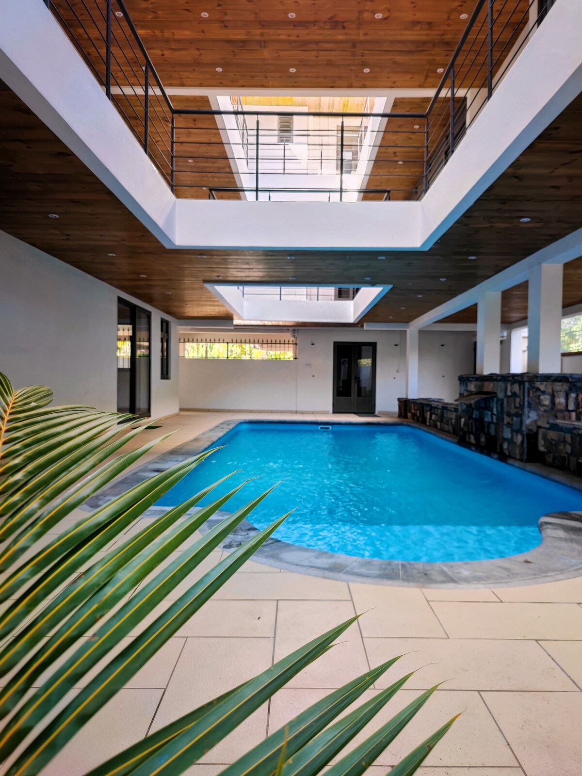 A crystal-clear pool is centrally positioned in a well-lit indoor area, surrounded by smooth tiles and natural wood accents. Large windows allow sunlight to penetrate, creating a lively atmosphere. Decorative plants border the pool area, enhancing the space's serene ambiance.