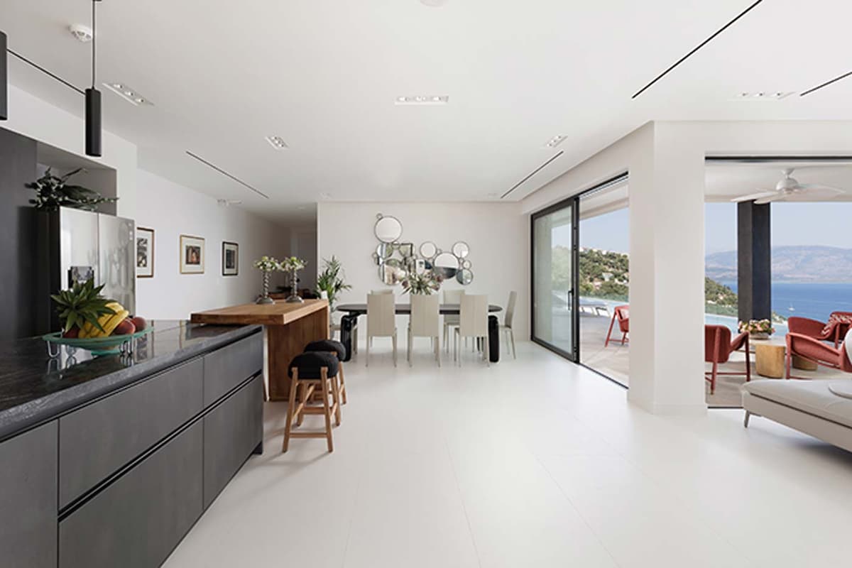 An open-plan living and dining area is visible, featuring a modern kitchen with a dark countertop and a wooden island. A dining table with chairs sits nearby, while large glass doors reveal an outdoor space with scenic views of the sea.