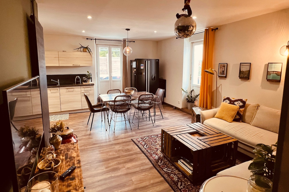 A contemporary living area showcases a comfortable seating arrangement and dining table. The space is illuminated by natural light from large windows. A kitchen area features modern cabinets and appliances. Decorative elements, including wall art and plants, add character to the inviting atmosphere.