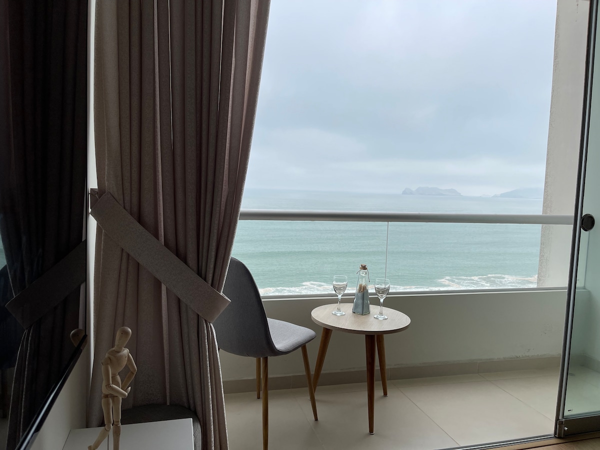 A balcony scene presents a table set with two glasses and a small bottle, with a clear view of the sea beyond. Soft curtains frame the space, while a minimalist chair provides a comfortable spot for relaxation. Gentle waves and distant land are visible in the background.