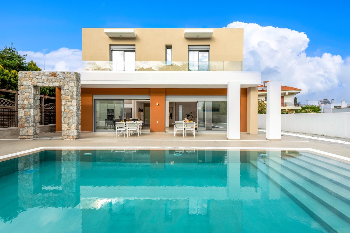 A modern villa with a stunning exterior is framed by a clear blue sky. A spacious private pool reflects the villa's design, while a patio area is furnished with a dining table and chairs, providing an inviting outdoor space for relaxation.