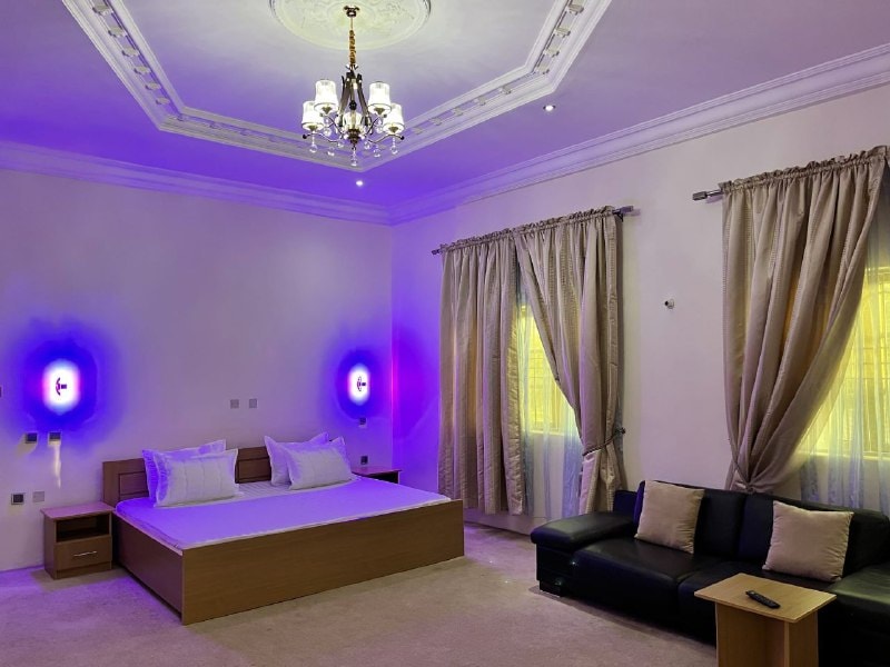 A spacious bedroom features soft, ambient lighting with purple hues. A large bed is positioned against the wall, accompanied by a dark sofa. Elegant curtains frame the windows, and a decorative chandelier enhances the room's ambiance.