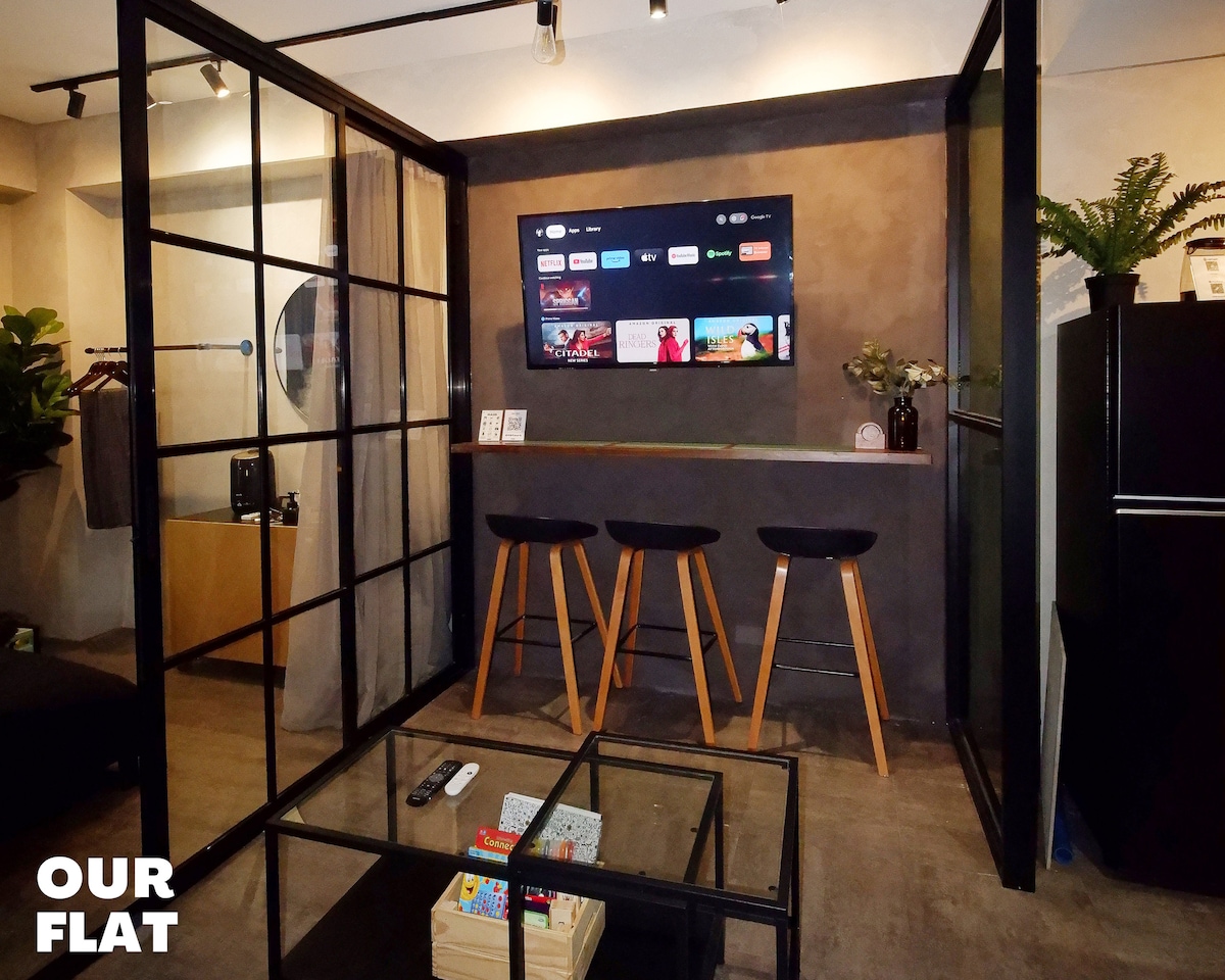 A modern living space features a sleek black-framed glass partition. A 50-inch 4K Smart TV displays various streaming options above a small hanging bar with three wooden stools. A low coffee table sits in front, accessorized with board games and a remote control.