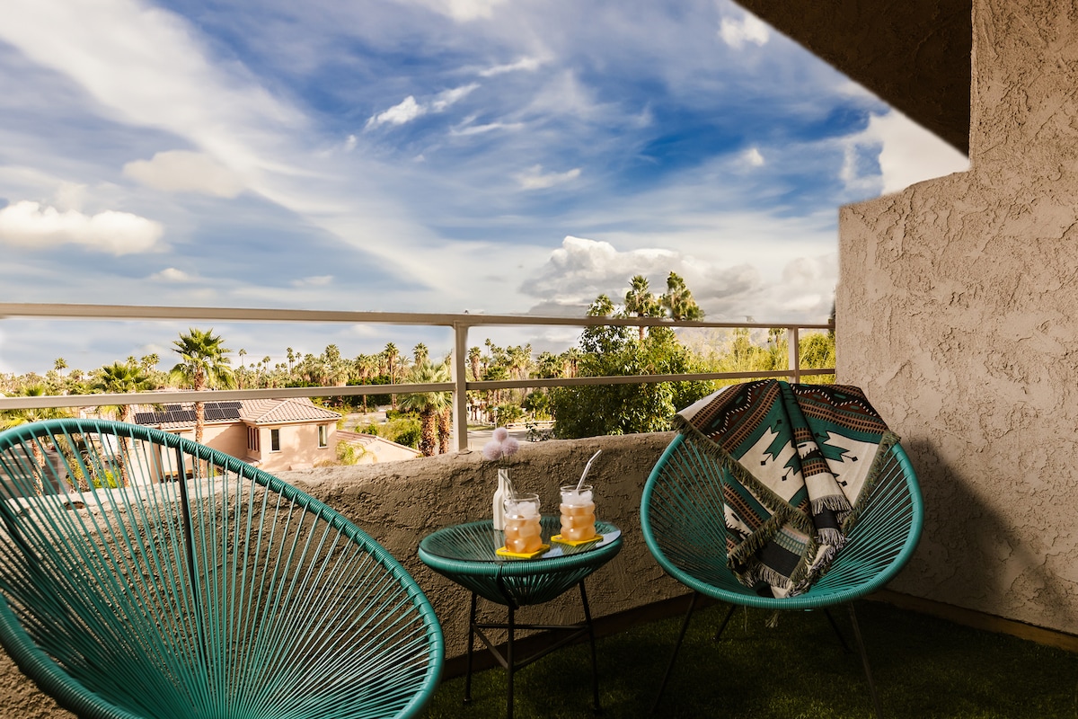 A cozy balcony setting features two vibrant turquoise chairs and a small table, adorned with two beverages. The backdrop showcases a clear blue sky with wispy clouds, and greenery from nearby palm trees is visible beyond the railing.