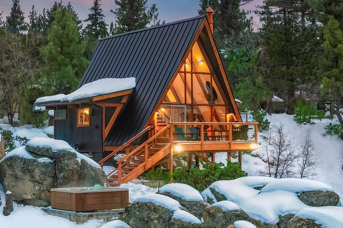 The A-Frame cabin is nestled among wintery landscapes, showcasing its distinctive architectural style and warm interior glow. Snow blankets the surrounding boulders and deck, where the hot tub is positioned nearby, offering an inviting contrast to the serene, icy scene.