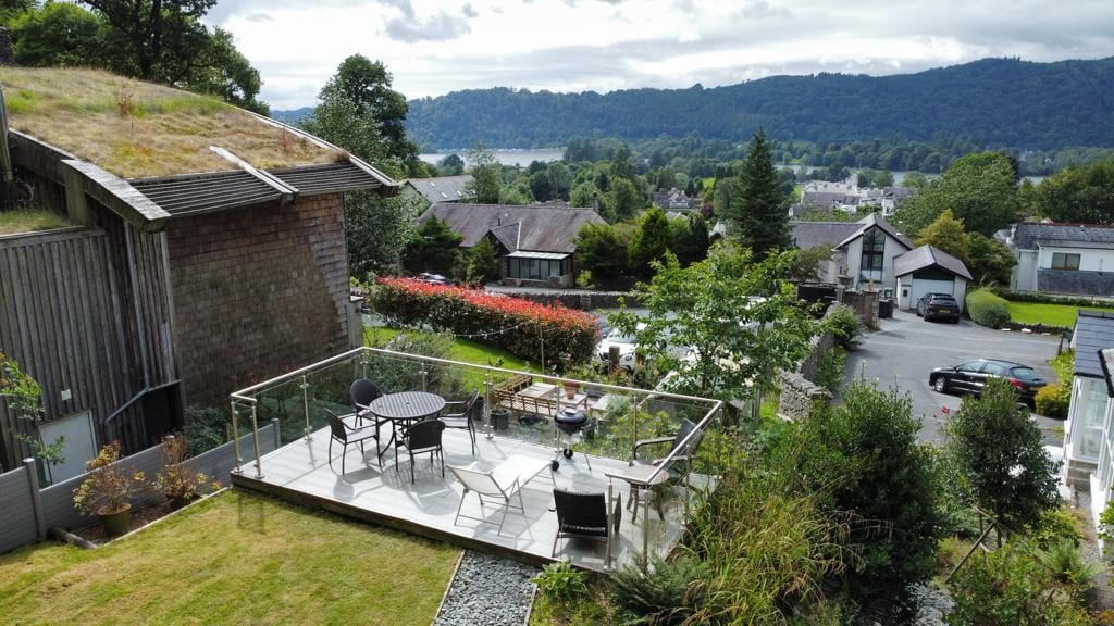 A private garden area features a raised deck with glass railings, equipped with a circular dining table and four chairs. The landscape includes lush greenery and views of the surrounding countryside, including distant hills and Lake Windermere, creating a serene outdoor setting.