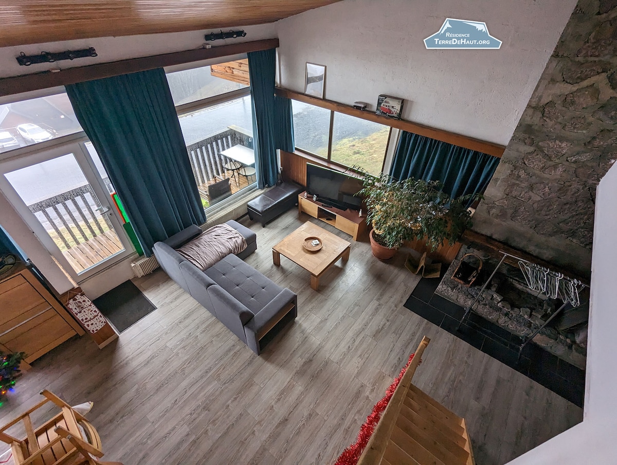 The living area features large windows allowing natural light to fill the space, offering views of the outdoors. A comfortable grey sofa is positioned in front of a wooden coffee table, with a television nearby. A decorative indoor plant adds a touch of greenery.