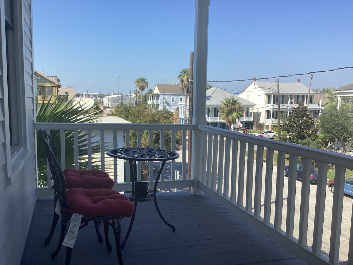 A charming front porch is adorned with a small round table and two black chairs, featuring red cushions. Palm trees and nearby houses are visible, with a clear blue sky enhancing the serene outdoor setting.