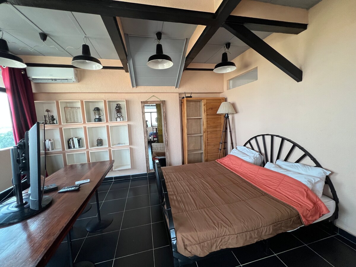 A well-lit room features a double bed with light brown bedding, complemented by white pillows. A large wooden desk is positioned nearby, alongside a flat-screen television. Dark tiles and exposed wooden beams add character to the space.