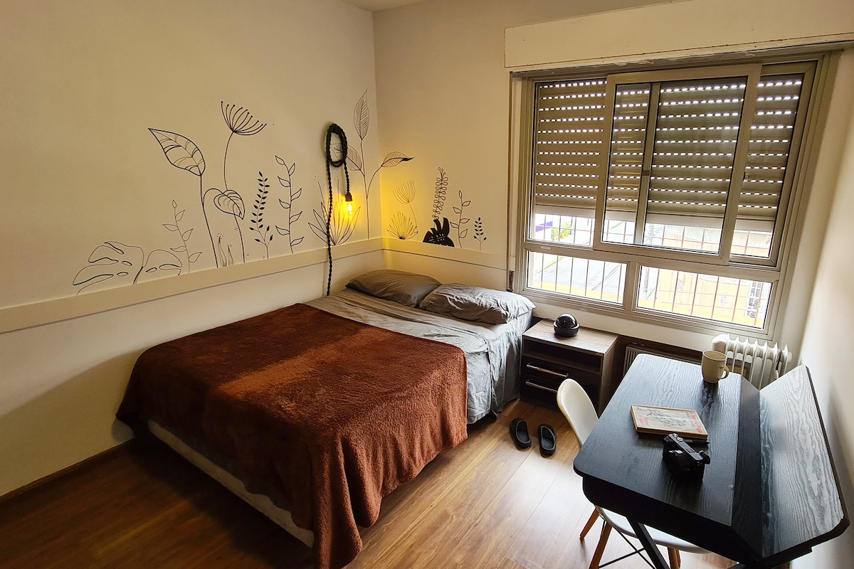A well-lit bedroom features a spacious bed covered with a brown blanket and gray pillows. Decorative illustrations adorn the wall above the bed. A compact desk and chair are positioned nearby, while a window with horizontal blinds allows natural light to filter in.