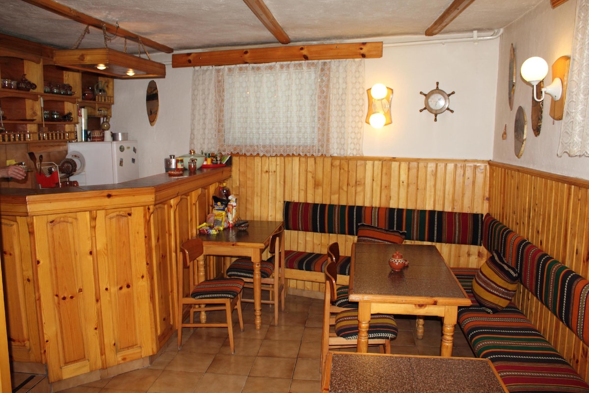 A communal area features wooden paneling and colorful striped seating. A countertop with kitchen essentials is visible, alongside a small dining table and chairs. Soft light illuminates the space, creating a cozy ambiance, while decorative elements add character to the room.
