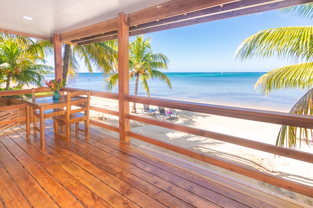 A wooden patio features a table and chairs positioned in front of large, open windows, providing an unobstructed view of the sandy beach and turquoise waters. Palm trees sway gently in the breeze, enhancing the tranquil outdoor setting.