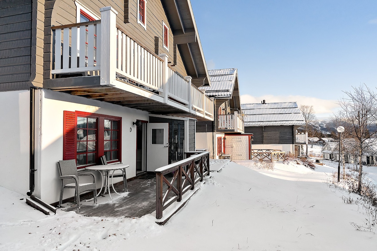 A welcoming exterior view of a ski-in/ski-out apartment surrounded by snow. The entrance features a covered patio with two gray chairs and a small table. Red shutters frame the windows, contrasting with the muted tones of the building. Nearby, other buildings provide a cozy atmosphere.