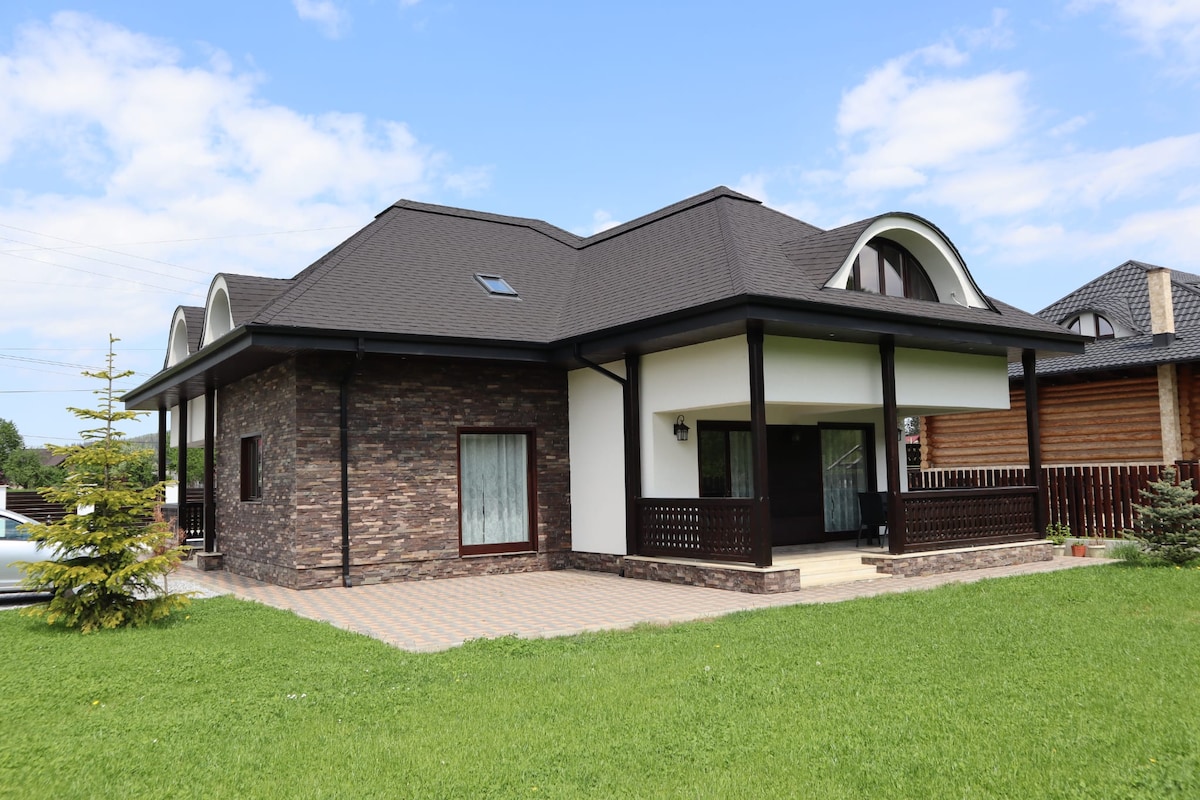 A modern house features a blend of stone and contemporary architecture. The exterior includes a spacious front porch with decorative railings. Lush green grass surrounds the building, while the visible roof showcases elegant curves. Clear windows enhance natural light across the façade.