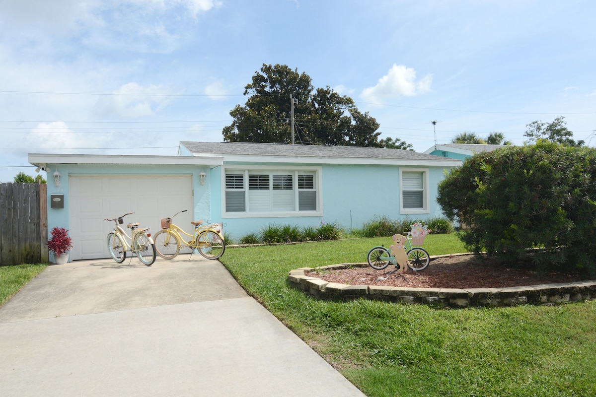 The exterior of the bungalow features a light blue facade with a well-maintained lawn. Two vintage bicycles, painted in yellow, are parked on the driveway, accompanied by a small pink children's bicycle. Decorative landscaping includes a shrub and mulch area.