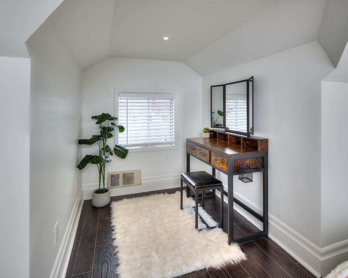 A well-appointed loft space is featured, designed for comfort and functionality. A wooden vanity table with a mirror sits against a sloped wall, accompanied by a black seating surface. A small potted plant adds a touch of greenery, while soft white carpeting enhances the inviting atmosphere.