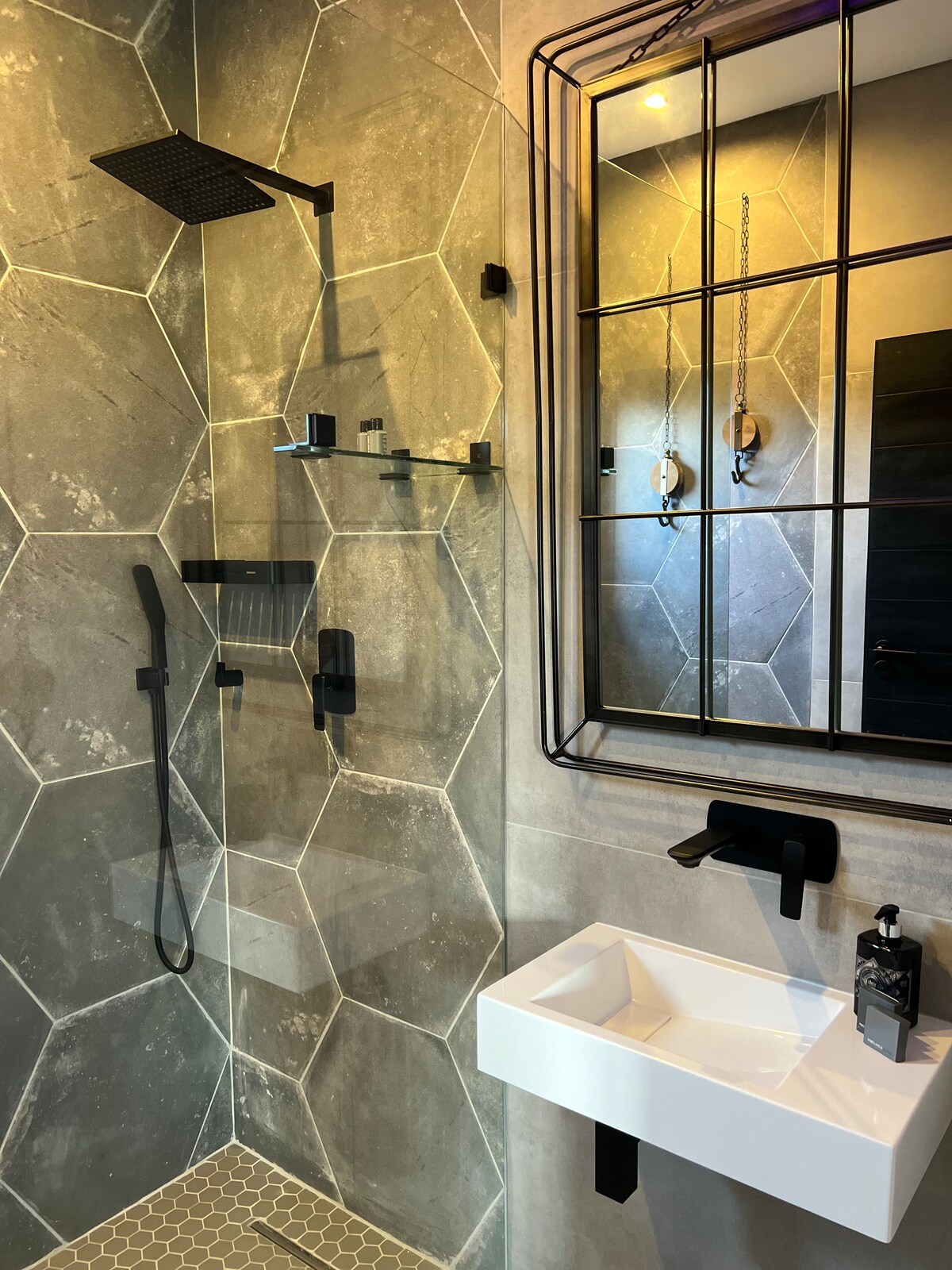 A modern shower area features geometric tile patterns on the walls. A sleek showerhead is mounted above, with a handheld option on the wall nearby. A compact sink with a minimalist design complements a large, framed mirror. Essential toiletries are neatly arranged.