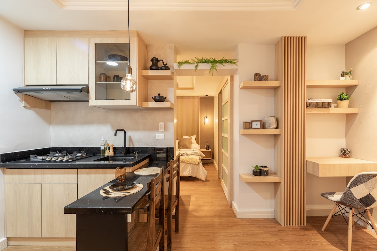 A modern kitchen is featured with wooden cabinetry and black countertops. Equipped with a stovetop and sink, it is complemented by a dining area with four wooden chairs. Shelving units along the walls display decorative items, while a glimpse of a cozy sleeping area is visible in the background.