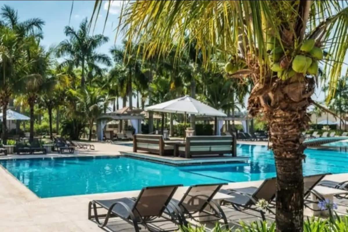 A swimming pool is surrounded by lush palm trees and lounge chairs. A shaded seating area with a canopy is positioned near the pool, enhancing relaxation options. Clear blue water reflects the sunlight, inviting leisurely moments under the warm sky.