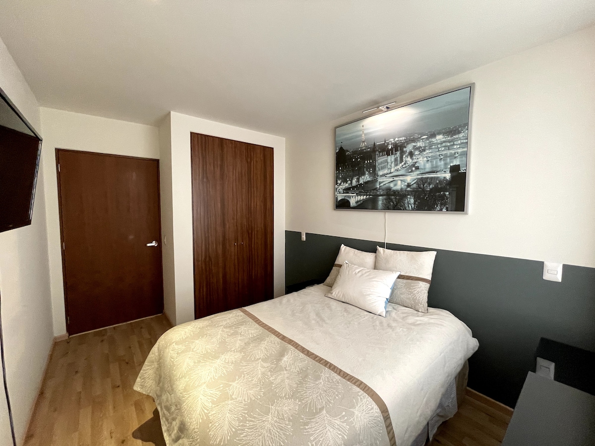 A sleeping area features a spacious bed with neutral bedding and a decorative throw. The wall above the bed displays a black and white photograph, while a wooden wardrobe provides storage. A flat-screen television is mounted on the opposite wall, enhancing functionality.