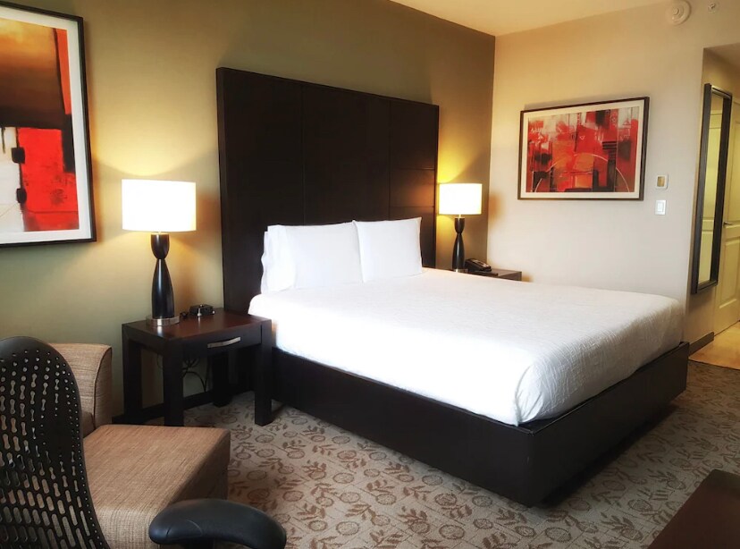 A modern guest room features a spacious bed with crisp white linens set against a dark wood headboard. Two contemporary lamps provide soft lighting next to the bed, complemented by a large piece of abstract art on the wall. A small armchair and a wooden table complete the setting.