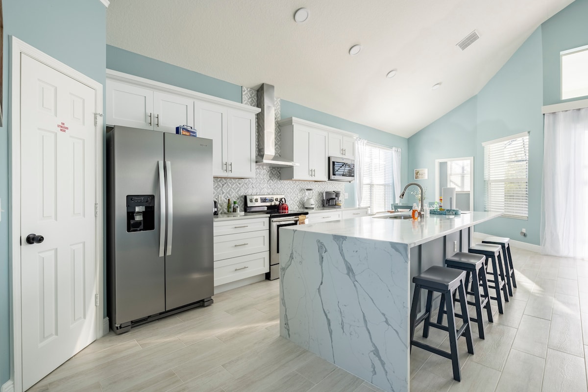 A modern kitchen features sleek white cabinetry and a spacious island with a marble-patterned countertop. Stainless steel appliances, including a refrigerator and oven, are complemented by a tiled backsplash. Four black bar stools are arranged around the island, with natural light pouring in from large windows.