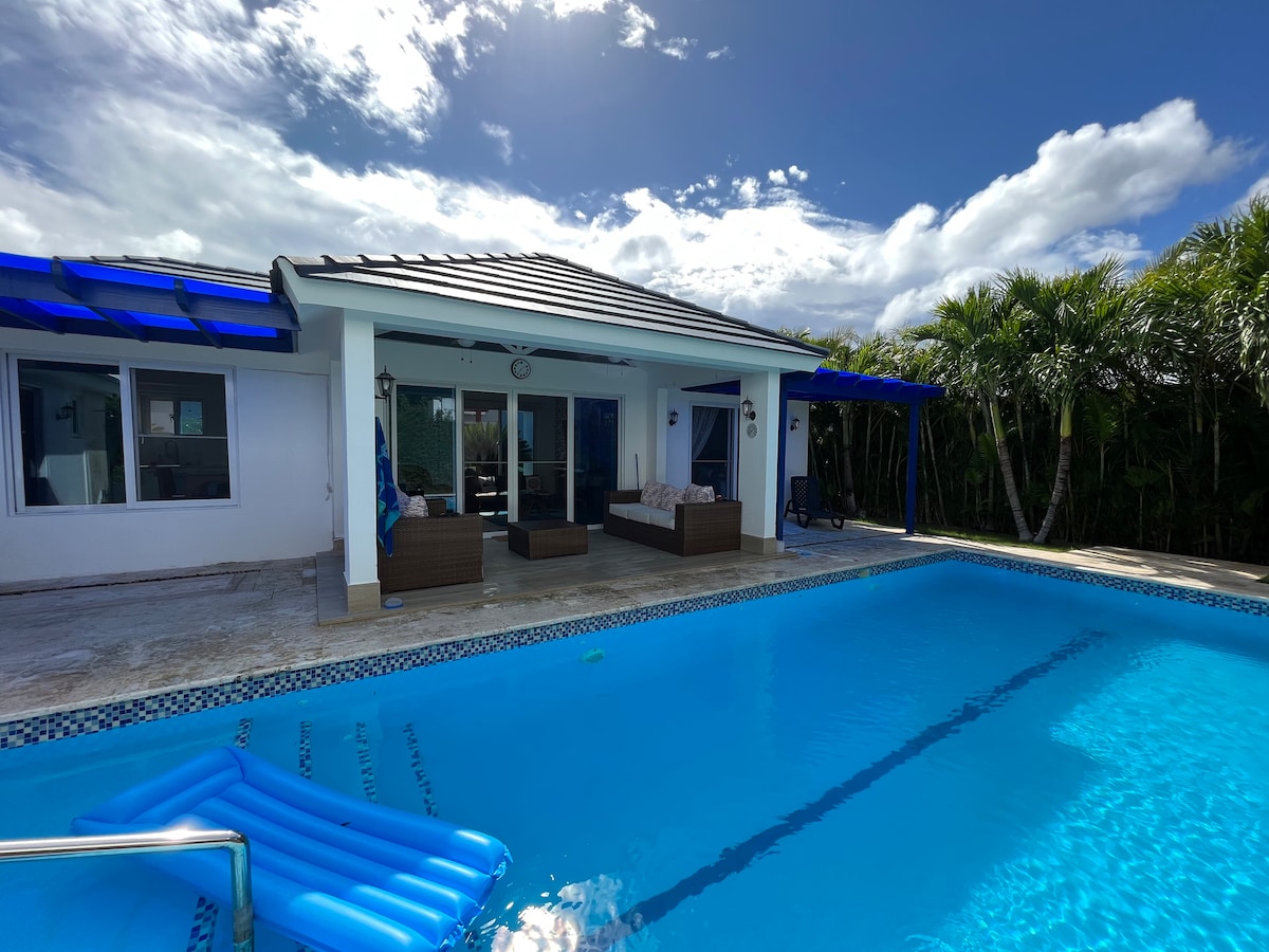 A modern villa with a spacious pool area is presented, featuring a large swimming pool and comfortable outdoor seating. The house is framed by lush greenery and boasts large windows allowing natural light. A bright blue sky with scattered clouds enhances the serene atmosphere.