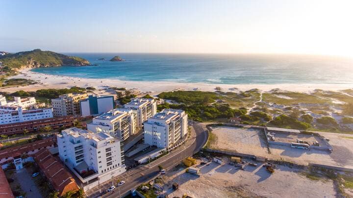 A panoramic view of the coastline reveals the sandy beach meeting the ocean waters. Nearby buildings line the shore, contrasted by green dunes. The light of the setting sun casts a warm glow over the scene, enhancing the tranquil atmosphere of the area.