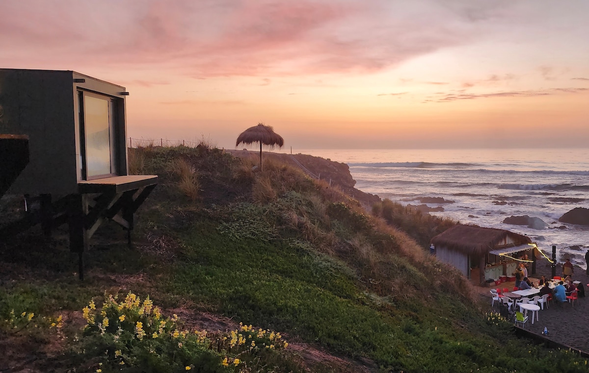 A hillside view captures a cabin with large windows, overlooking the ocean at sunset. A thatched umbrella provides shade nearby, while a gathering area with tables and chairs is seen in the background, surrounded by lush greenery and contrasting rocky coastline.