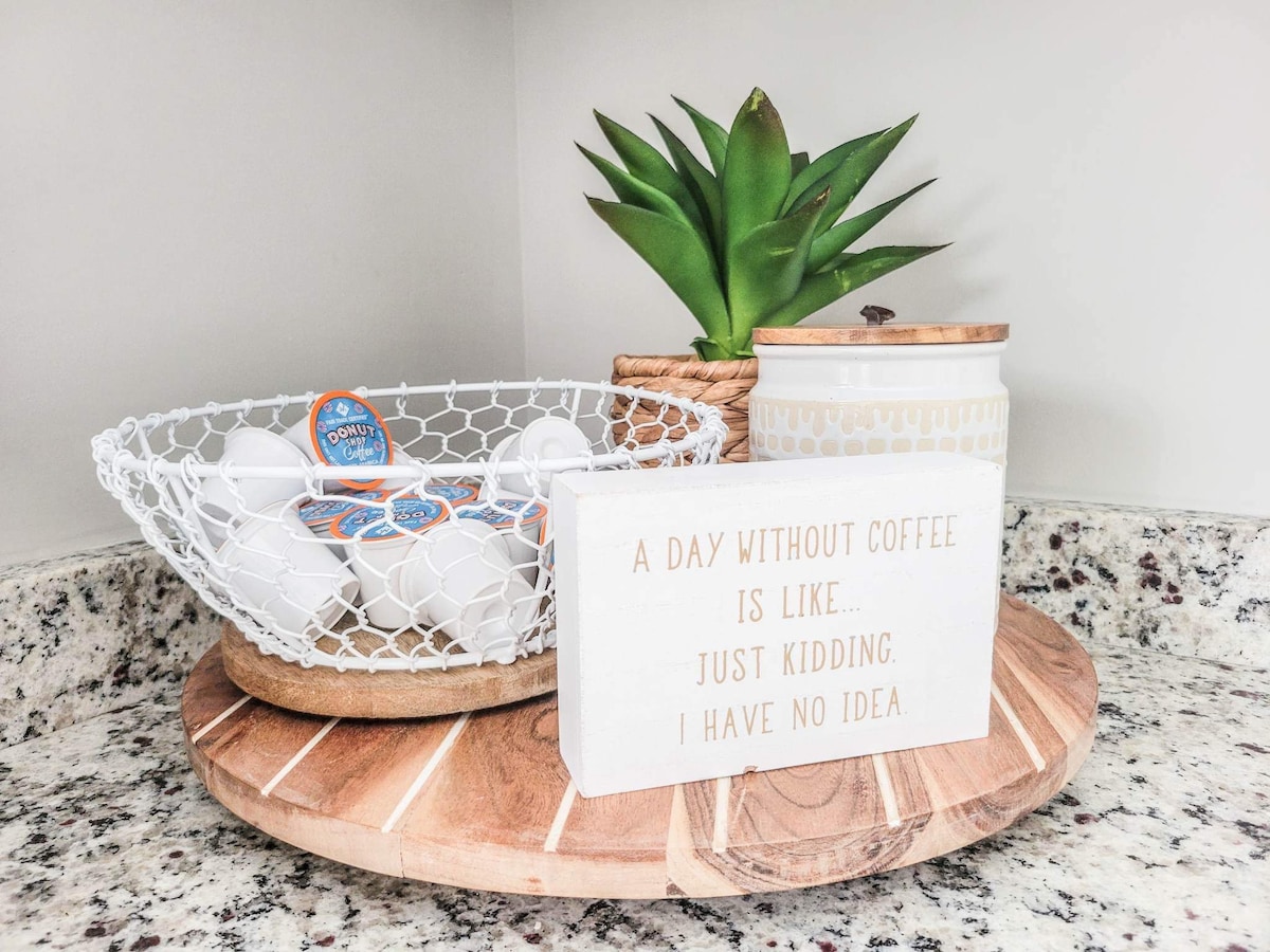 A decorative kitchen counter arrangement features a round wooden tray displaying a wire basket filled with coffee pods. A small potted succulent and a ceramic container add greenery, while a sign with a humorous coffee-related saying rests on the tray.