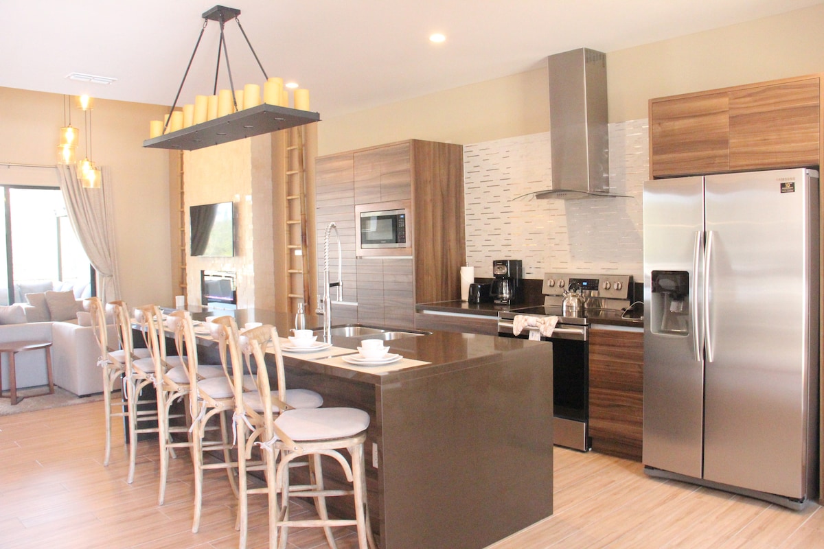 A modern kitchen features a central island with seating for six. Stainless steel appliances are complemented by wooden cabinetry. Natural light filters through large windows, illuminating the space. Decorative pendant lights hang above the island, and a cozy seating area is visible in the background.