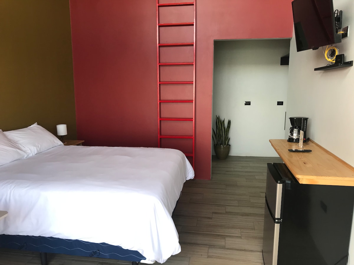 A spacious sleeping area features a large bed with crisp white linens. A ladder leads to a higher sleeping space, while a small kitchenette with a coffee maker and a mini fridge is visible. The walls are painted in warm tones, contributing to a cozy ambiance.