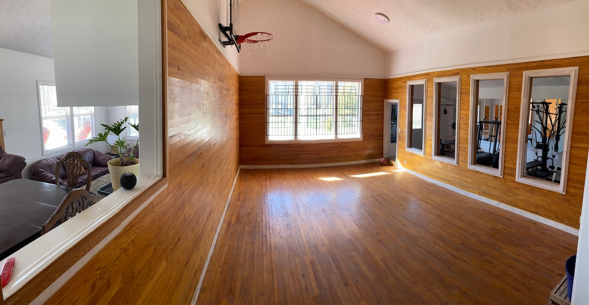 A spacious room is highlighted by polished wooden floors and high ceilings. Basketball equipment is mounted on the wall, and large windows allow natural light to fill the area. Mirrors are positioned along one side, reflecting the open space.