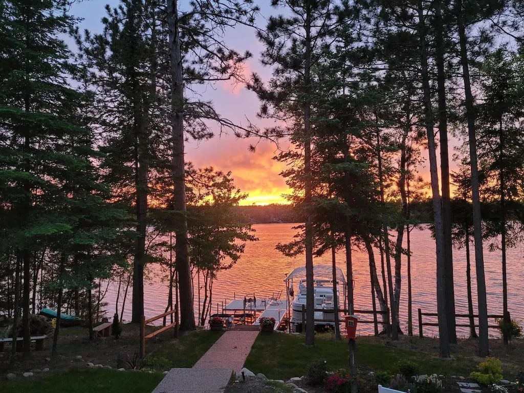 A scenic view of a tranquil lake at sunset is framed by tall trees. The colorful sky reflects on the water, while a dock with a boat is visible in the foreground. A stone pathway leads toward the lakeshore, enhancing the natural beauty of the setting.