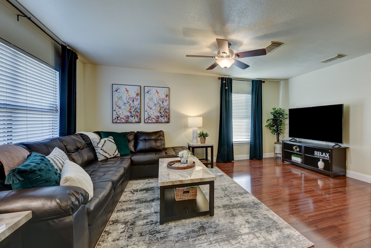 A spacious living area showcases a sectional leather sofa arranged around a coffee table with decorative items. Two abstract paintings adorn the wall, while a television is positioned on a console unit. Natural light enters through the window draped with dark curtains.
