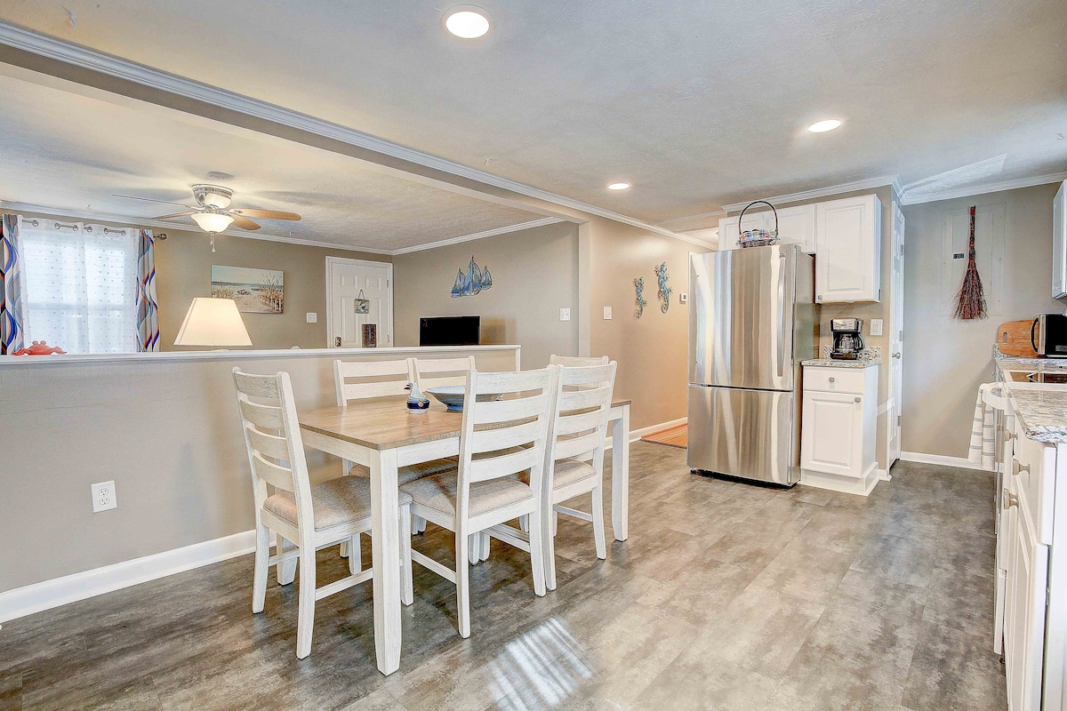 An open kitchen and dining area is depicted, featuring a wooden dining table surrounded by six white chairs. A stainless steel refrigerator stands alongside white cabinetry, with a ceiling fan providing overhead lighting. Light-colored walls enhance the spaciousness of the room.